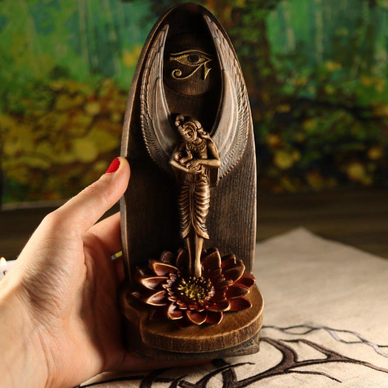 K&ouml;nnte beinhalten: Eine braune und goldfarbene dekorative Statue mit einer Engelsfigur mit ausgebreiteten Fl&uuml;geln, die auf einer Lotusblume steht. Die Statue hat ein Horusauge-Symbol und ist vor einem dunklen Holzgrund platziert.