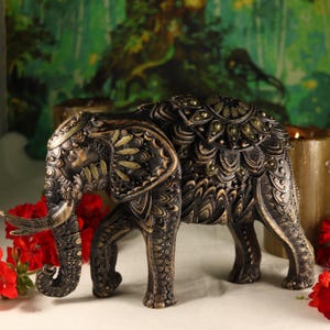 May include: A black and gold elephant figurine with intricate carvings and green gemstone accents. The elephant is standing on a white surface.