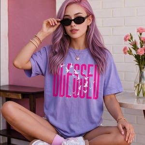 Puede incluir: Una camiseta de manga corta de color morado claro con la palabra BLESSED impresa en letras mayúsculas de color rosa intenso en el pecho, con pequeñas siluetas de aves blancas intercaladas en el texto, creando un diseño gráfico frontal.