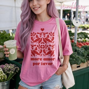 Puede incluir: Una camiseta rosa con un gráfico rojo de flores estilizadas, un pequeño corazón y el texto more amor por favor impreso en un marco rectangular. La camiseta tiene mangas cortas y cuello redondo, combinada con pantalones cortos de mezclilla.