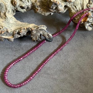 Op de afbeelding: Een enkelstrengs ketting bestaande uit kleine, gefacetteerde, diep magentakleurige kralen. De ketting is voorzien van een metalen sluiting aan het uiteinde van de streng, waarbij de kralen in een doorlopende, gelijkmatige cirkelvorm liggen.
