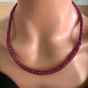 May include: A necklace composed of small, faceted, deep red gemstones. The necklace is draped around a person's neck, showcasing the vibrant color and the symmetrical arrangement of the beads. The gemstones appear to be polished and reflect light.