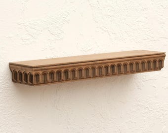 May include: A rectangular, wooden shelf with a decorative, arched design along the front edge. The shelf is a warm, medium brown color and appears to be mounted on a textured, off-white wall. The shelf is approximately 30 inches long.