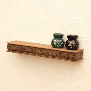 May include: A carved wooden shelf displaying two ceramic oil diffusers. One is dark green, the other black. The shelf has detailed carvings and is a warm brown colour. The shelf is mounted on a textured white wall.