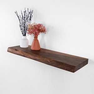 Può includere: Una mensola in legno marrone scuro, con bordo vivo, montata su una parete bianca. Due piccoli vasi con fiori secchi sono appoggiati sulla mensola. Un vaso è bianco e scanalato, l'altro è color terracotta.