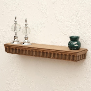 Peut inclure: Une étagère en bois avec un bord décoratif, supportant deux bougeoirs en verre transparent et un petit diffuseur d'huiles essentielles en céramique verte. L'étagère est fixée à un mur texturé blanc cassé.