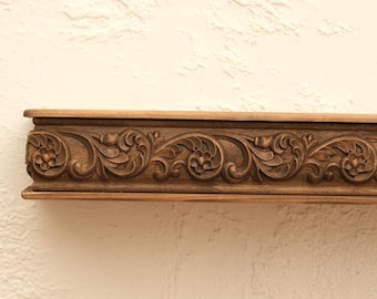 May include: A section of a wooden decorative molding with intricate floral and leaf carvings. The molding is a warm brown color and features a repeating pattern of stylized flowers and swirling leaves. The wood has a natural grain.