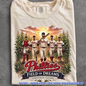 Peut inclure: Un t-shirt couleur crème avec une illustration de cinq joueurs de baseball en uniforme blanc dans un champ de maïs au coucher du soleil. Le texte indique Phillies, Field of Dreams et Cornfield Classic. Étiquette Comfort Colors au col.