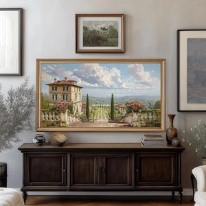 May include: A large framed painting depicts a villa with a garden, set against a backdrop of a blue sky with fluffy clouds. The painting is in a gold frame, and is hung above a dark wood cabinet.