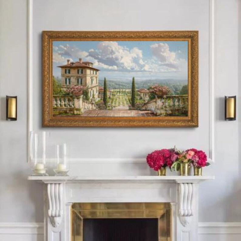 May include: A framed landscape painting depicts a villa with a garden, under a cloudy blue sky. The painting has a gold ornate frame. Below the painting is a white mantel with candles and floral arrangements in gold vases. Two gold sconces are on either side of the painting.