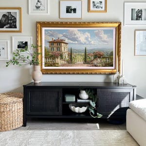 May include: A large framed landscape painting with a gold frame hangs above a black cabinet. The painting depicts a villa with a garden and a cloudy sky. The cabinet has woven door panels and is decorated with a vase, bowl, and greenery.