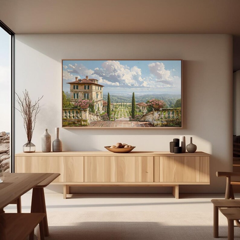 May include: A framed landscape painting depicts a villa with a terraced garden under a cloudy sky. The painting hangs above a light wood cabinet with decorative vases and a wooden bowl. The room has a modern design with natural light.