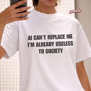 Może przedstawiać: Biała koszulka z czarnym napisem: "AI CAN'T REPLACE ME I'M ALREADY USELESS TO SOCIETY". Słowo "white" jest napisane kursywą w prawym górnym rogu. Koszulka jest w stylu casual, z krótkim rękawem.