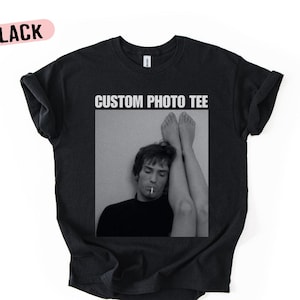 Può includere: T-shirt nera con stampa fotografica in bianco e nero. La foto mostra una persona con una sigaretta e gambe in aria. Il testo "CUSTOM PHOTO TEE" è stampato sopra la foto. La parola "BLACK" è stampata su un ovale rosa.