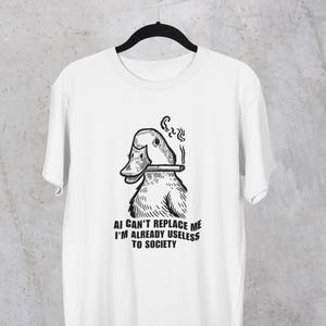 May include: White t-shirt with a black and white graphic of a smoking duck. The text below the duck reads "AI CAN'T REPLACE ME I'M ALREADY USELESS TO SOCIETY."