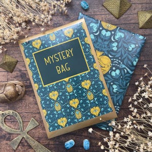 Könnte beinhalten: Draufsicht auf eine „MYSTERY BAG“ mit einem teal-goldenen Design, neben einer gemusterten Verpackung. Die Tasche hat einen dunkelblauen Hintergrund mit goldenen Akzenten und dem Text „MYSTERY BAG“. Dekorative Elemente sind goldene Pyramiden und ein Ankh.