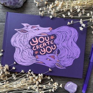 Könnte beinhalten: Ein lila Notizbuch mit einer Wolfsillustration und dem Text "YOU CREATE YOU" in einer verspielten Schriftart. Das Kunstwerk zeigt zwei Wölfe mit fließendem lila Haar. Das Notizbuch ist von Trockenblumen und einem lila Stift umgeben.