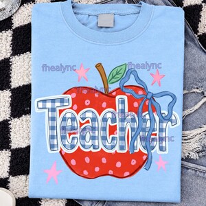 May include: A light blue t-shirt featuring a graphic of a red apple with white polka dots, a brown stem, and a green leaf. The word Teacher is printed in a blue and white checkered pattern across the apple, accented by a blue ribbon and pink stars.