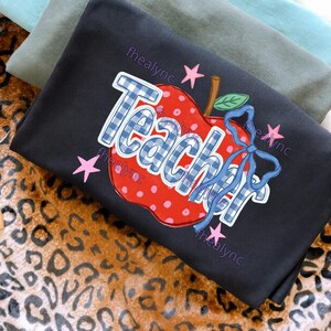May include: A black t-shirt featuring a red apple graphic with white polka dots and a blue gingham pattern on the word Teacher. The design includes a blue ribbon tied around the apple stem, green leaves, and small pink stars scattered around the text.