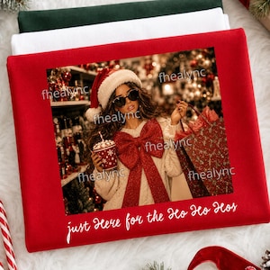 May include: A red sweatshirt featuring a printed graphic of a person wearing a Santa hat, sunglasses, and a cream-colored sweater with a large red bow. The person holds a red mug. The text "just Here for the Ho Ho Hos" is printed in white.