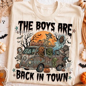 May include: A cream-colored t-shirt featuring a graphic of a vintage van filled with Halloween characters, including a skeleton, a werewolf, and a ghost. The text reads THE BOYS ARE BACK IN TOWN, surrounded by pumpkins, bats, and a large orange moon.