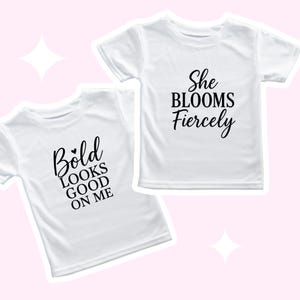 May include: Two white t-shirts with black text. One shirt says "Bold Looks Good On Me" with a small heart above the word "Bold". The other shirt says "She Blooms Fiercely".