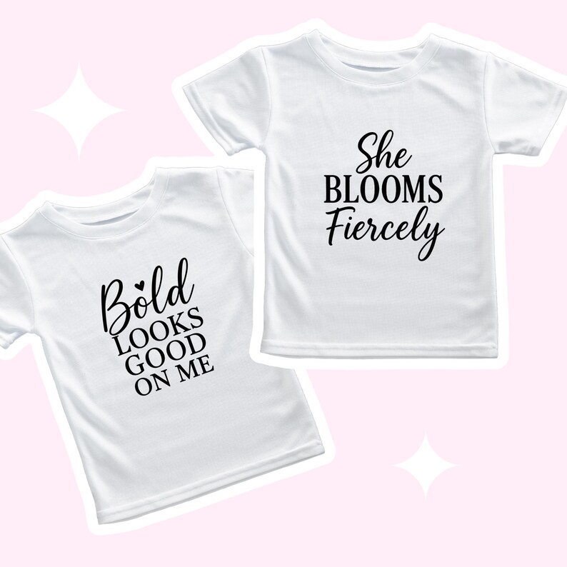 May include: Two white t-shirts with black text. One shirt says "Bold Looks Good On Me" with a small heart above the word "Bold". The other shirt says "She Blooms Fiercely".