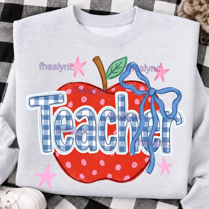 May include: A light gray crewneck sweatshirt featuring a large red apple graphic with pink polka dots, a green leaf, and a blue ribbon bow. The word Teacher is printed across the apple in a blue and white gingham check pattern with pink stars.