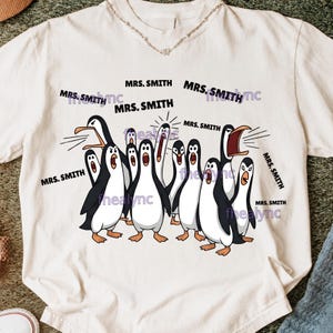 May include: A white t-shirt featuring a cartoon illustration of a group of penguins with open beaks. The text "MRS. SMITH" is printed around the penguins. The shirt has a crew neck and short sleeves.