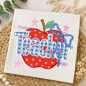 May include: A white t-shirt featuring a red apple graphic with pink polka dots, a brown stem, and a green leaf. The word Teacher is printed in a blue and white gingham pattern across the apple, accented by a blue ribbon and four pink stars.