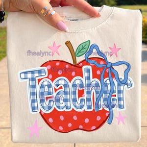May include: A cream-colored t-shirt featuring a large red apple graphic with pink polka dots, a brown stem, and a green leaf. The word Teacher is printed in blue and white gingham letters, accented by a blue ribbon bow and small pink stars.