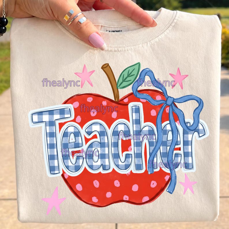 May include: A cream-colored t-shirt featuring a large red apple graphic with pink polka dots, a brown stem, and a green leaf. The word Teacher is printed in blue and white gingham letters, accented by a blue ribbon bow and small pink stars.