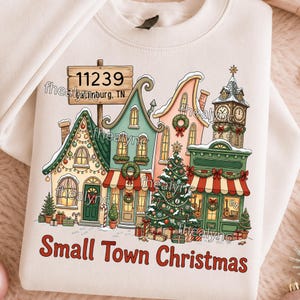 Puede incluir: Una sudadera color crema con un gráfico de una escena de pueblo festivo, edificios cubiertos de nieve, un árbol de Navidad decorado y un letrero de madera que dice 11239 Gatlinburg, TN. El texto Small Town Christmas aparece en rojo abajo.