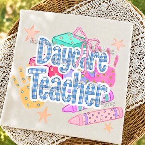 May include: A white t-shirt featuring a graphic print with the text Daycare Teacher in blue and white gingham letters. The design includes a teal gift box with a pink bow, a yellow handprint, pink crayons with polka dots, and light orange stars.