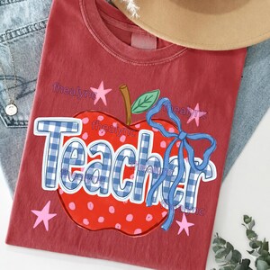 May include: A brick red t-shirt featuring a large red apple graphic with white polka dots, a green leaf, and a blue gingham bow. The word Teacher is printed in large blue and white gingham letters across the apple, surrounded by small pink stars.