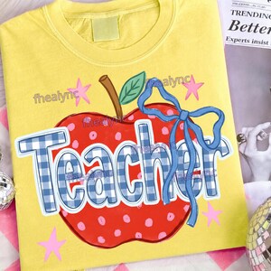 May include: A bright yellow t-shirt featuring a large red apple graphic with pink polka dots, a green leaf, and a blue ribbon bow. The word Teacher is printed across the apple in a blue and white gingham pattern. Pink stars surround the apple design.