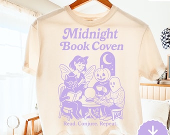 May include: Cream-coloured t-shirt with a purple graphic. The graphic features the text "Midnight Book Coven" and the phrase "Read. Conjure. Repeat." The design shows a cat, fairy, pumpkin, and ghost reading books around a table.