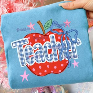 May include: A light blue sweatshirt featuring a large red apple graphic with white polka dots, a brown stem, and a green leaf. The word Teacher is printed in a blue and white gingham pattern, accented by a blue ribbon bow and small pink stars.