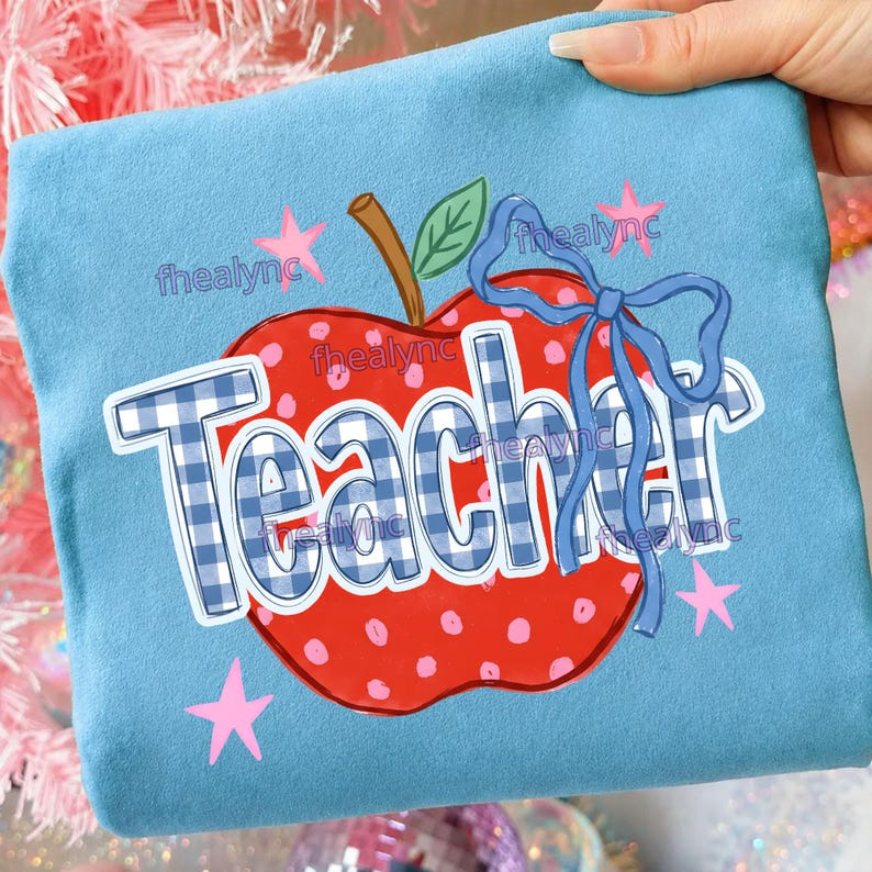 May include: A light blue sweatshirt featuring a large red apple graphic with white polka dots, a brown stem, and a green leaf. The word Teacher is printed in a blue and white gingham pattern, accented by a blue ribbon bow and small pink stars.