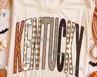 May include: A cream-coloured t-shirt featuring the word KENTUCKY in large, stylised letters. Each letter has a unique pattern, including orange and brown checkers, pumpkins, plaid, leaves, and polka dots. The design is centred on the front.