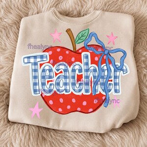 May include: A beige crewneck sweatshirt featuring a large red apple graphic with pink polka dots, a brown stem, and a green leaf. The word Teacher is printed in blue and white gingham letters, accented by a blue ribbon bow and pink stars.
