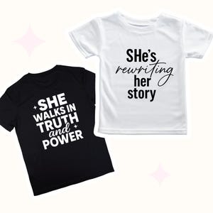 May include: Two short-sleeved t-shirts, one black and one white. The black shirt has the text "SHE WALKS IN TRUTH and POWER" in white. The white shirt says "She's rewriting her story" in black script. Both shirts are on a light pink background.