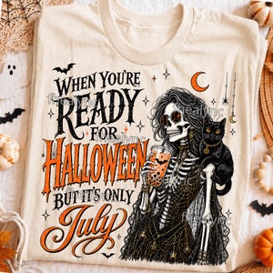 May include: A cream-colored t-shirt featuring a graphic of a skeleton wearing sunglasses and holding an orange drink, with a black cat on its shoulder. The text reads: When you're ready for Halloween but it's only July. Includes bats, stars, and a moon.