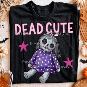 May include: A black t-shirt featuring the text DEAD CUTE in pink letters above a graphic of a grey doll with button eyes and stitched mouth, wearing a purple dress with white polka dots, accented by two pink stars on the sides of the doll.