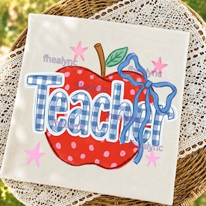 May include: A square off-white fabric item featuring a red apple graphic with pink polka dots, a brown stem, and a green leaf. The word Teacher is printed in a blue and white gingham pattern, accented by a blue ribbon bow and small pink stars.