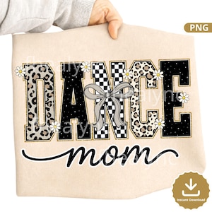 May include: A beige fabric item featuring the words "DANCE mom" in a decorative design. The word "DANCE" is in a bold, block letter style with a gold outline, incorporating leopard print, checkered patterns, and black. A gray bow and daisy accents are included.