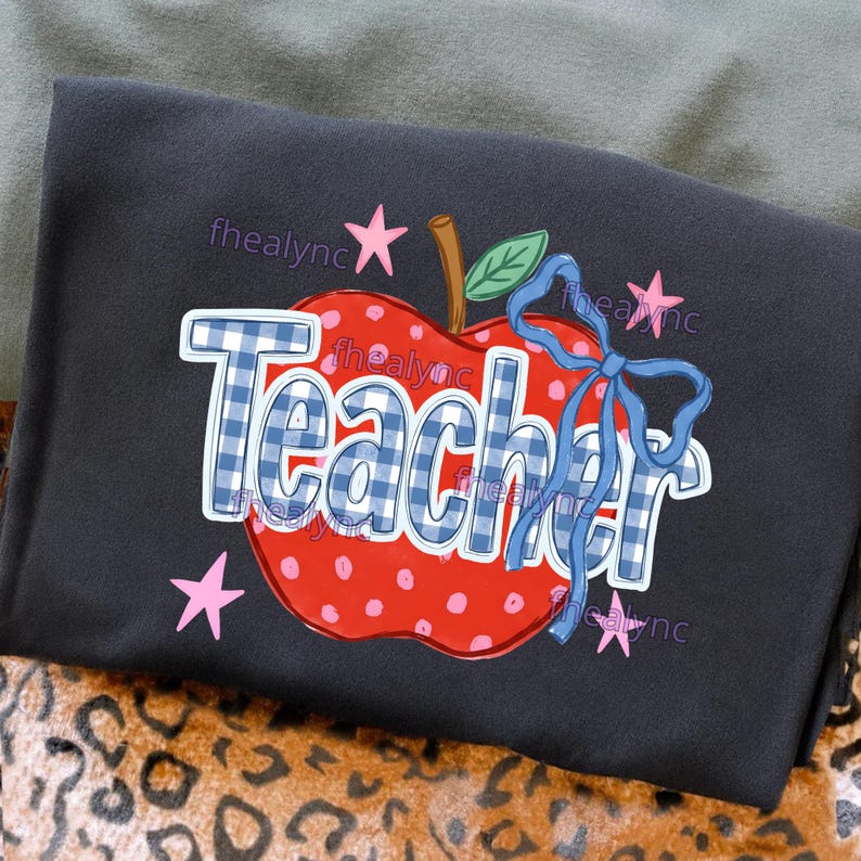 May include: A black t-shirt featuring a graphic of a red apple with white polka dots. The word Teacher is written in a blue and white gingham pattern across the apple. A blue ribbon is tied to the apple stem, and pink stars surround the design.