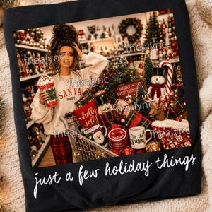 May include: A black t-shirt featuring a graphic of a person in a beige sweatshirt with the text SANTA BABY, holding a festive cup. The graphic includes a shopping cart filled with holiday decor, a nutcracker, and a mug labeled tis the season. The text just a few holiday things is printed below the image.
