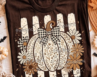 May include: A dark brown t-shirt featuring a central graphic of a white pumpkin decorated with intricate brown floral line art. The pumpkin is topped with a black and white buffalo plaid bow and surrounded by various brown and white daisy flowers.