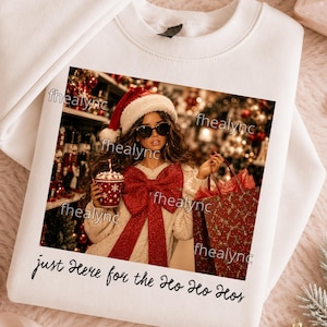 May include: A white crewneck sweatshirt featuring a graphic print of a person wearing a Santa hat and sunglasses, holding a red cup and a red gift bag. The person wears a cream sweater with a large red bow. Text reads: just Here for the Ho Ho Hos.
